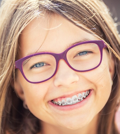 A young girl wearing purple glasses smiles while showing off her new braces put into place by her orthodontist in Plano